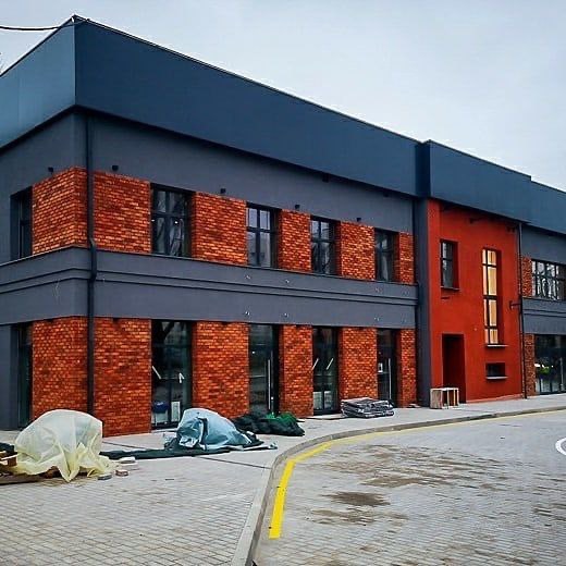 Dwukondygnacyjny budynek biurowy z szarą elewacją, fragmentami wykończonymi czerwoną cegłą i pomarańczową ścianą, widoczne materiały budowlane przed budynkiem.
