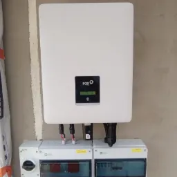 Biały inwerter marki FOX podłączony do dwóch skrzynek zabezpieczających na szarej ścianie wewnątrz budynku.