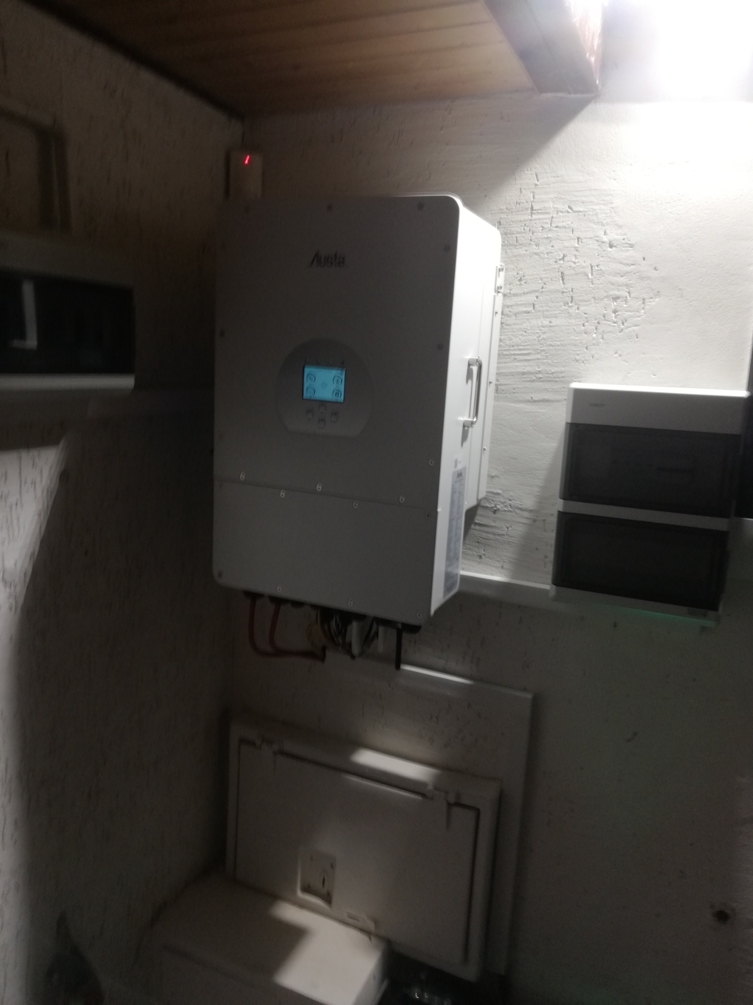 Biały inwerter fotowoltaiczny marki Austa zamontowany na szarej, chropowatej ścianie w pomieszczeniu technicznym, widoczne kable i skrzynka rozdzielcza pod spodem.