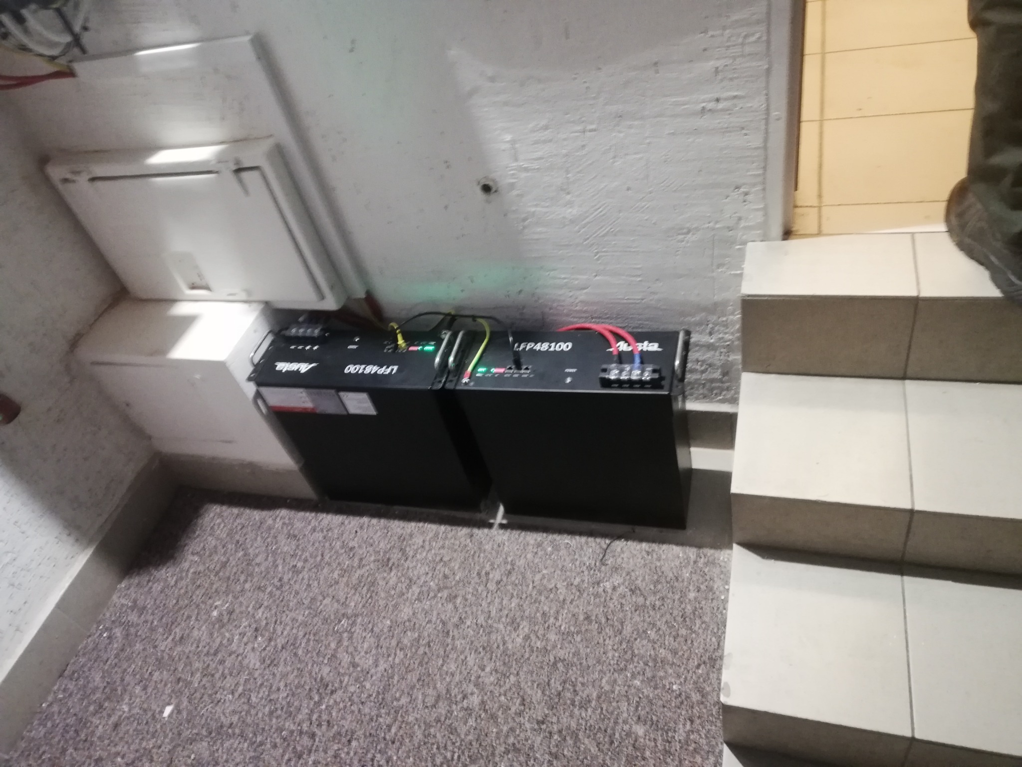 Dwa czarne moduły akumulatorowe LFP48100 marki Alsta, połączone kablami, umieszczone w pomieszczeniu z szarą wykładziną i białymi ścianami, widoczne z góry, obok schodów wyłożonych płytkami.