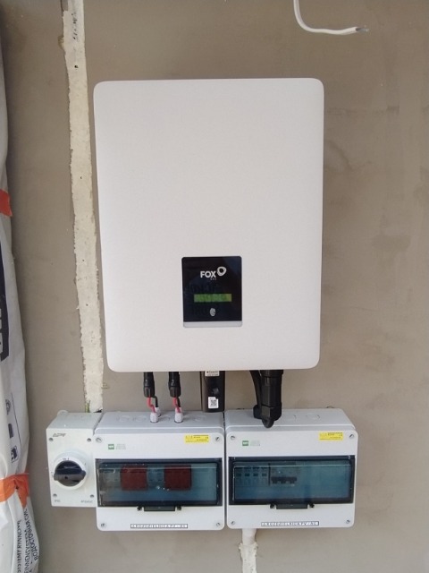 Biały inwerter marki FOX podłączony do dwóch skrzynek zabezpieczających na szarej ścianie wewnątrz budynku.