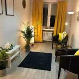 Jasny gabinet psychologiczny z żółtymi zasłonami, dywanem w kolorze granatowym, roślinami doniczkowymi i minimalistycznymi obrazami na ścianach. W tle widoczny grzejnik.