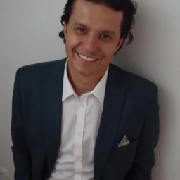 Uśmiechnięty mężczyzna w ciemnozielonym garniturze i białej koszuli, z kolorową poszetką w kieszeni, oparty o białą ścianę.