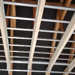 Konstrukcja sufitu podwieszanego z płyt kartonowo-gipsowych, widoczne profile metalowe i drewniane elementy więźby dachowej, czarna izolacja, drabina w rogu.