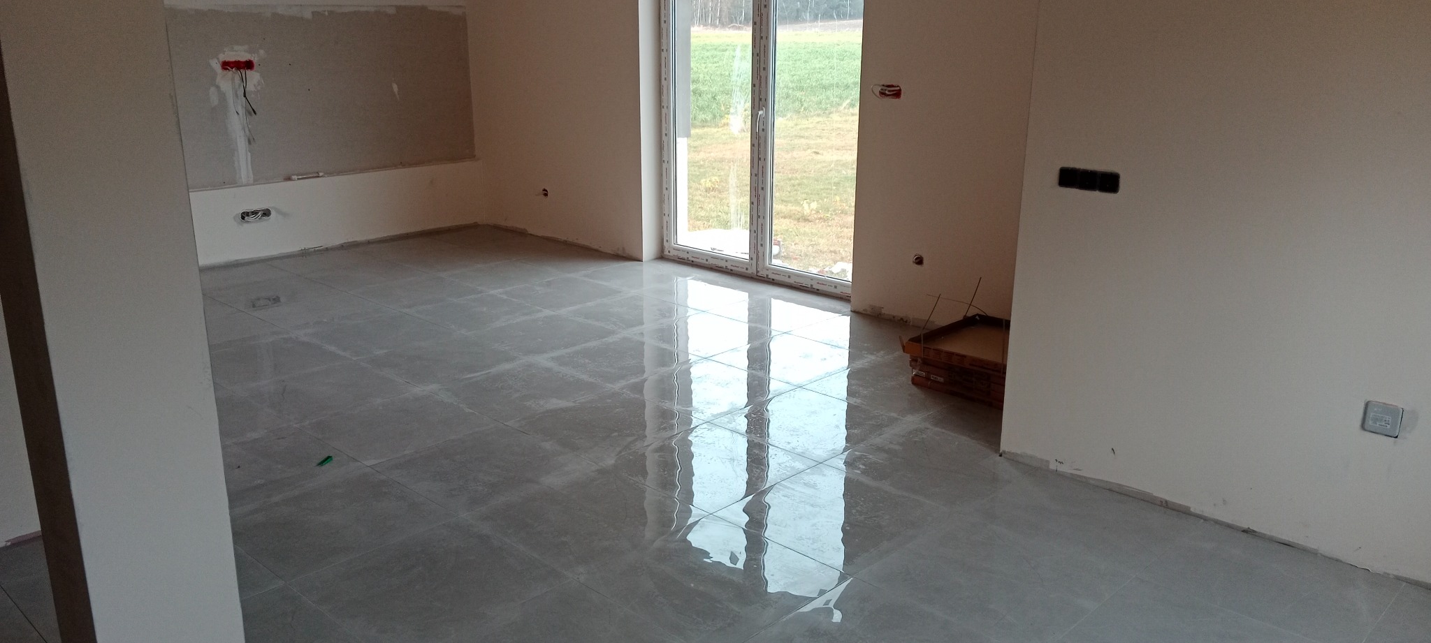 Szara, błyszcząca podłoga z płytek w nowym mieszkaniu, widoczne przygotowane ściany do dalszych prac wykończeniowych i okno z widokiem na zewnątrz.