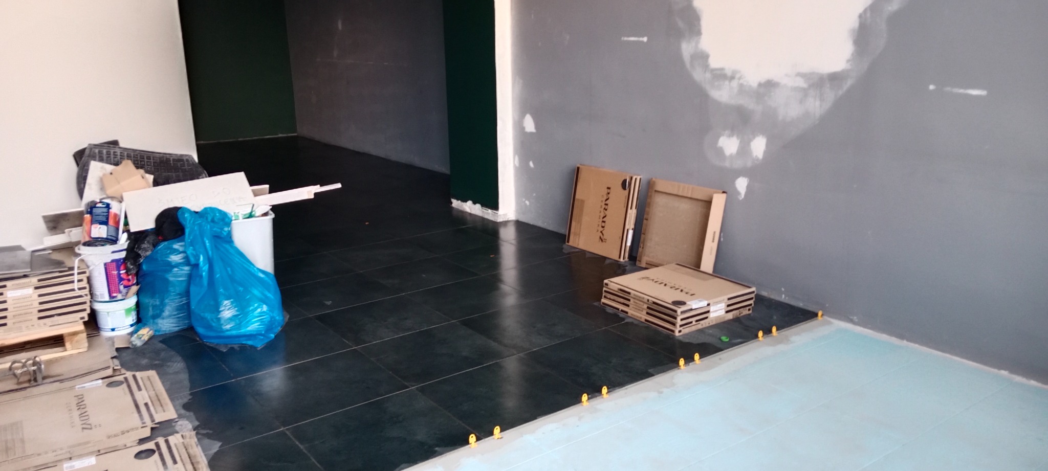 Fragment pomieszczenia w trakcie kładzenia ciemnych płytek podłogowych, widoczne opakowania po płytkach, worki z odpadami i poziomowanie płytek za pomocą żółtych krzyżyków.