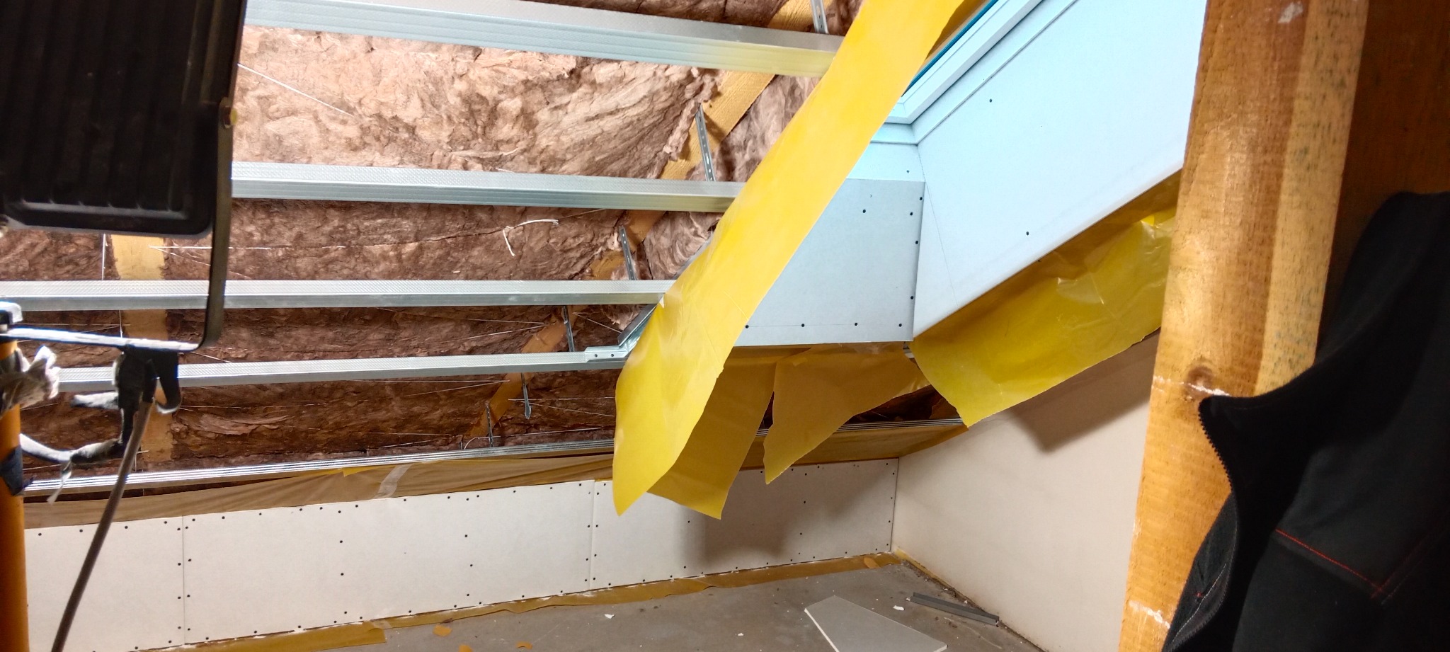 Montaż sufitu podwieszanego z płyt kartonowo-gipsowych, widoczna konstrukcja metalowa, izolacja z wełny mineralnej, zabezpieczenie folią malarską i fragment okna dachowego.