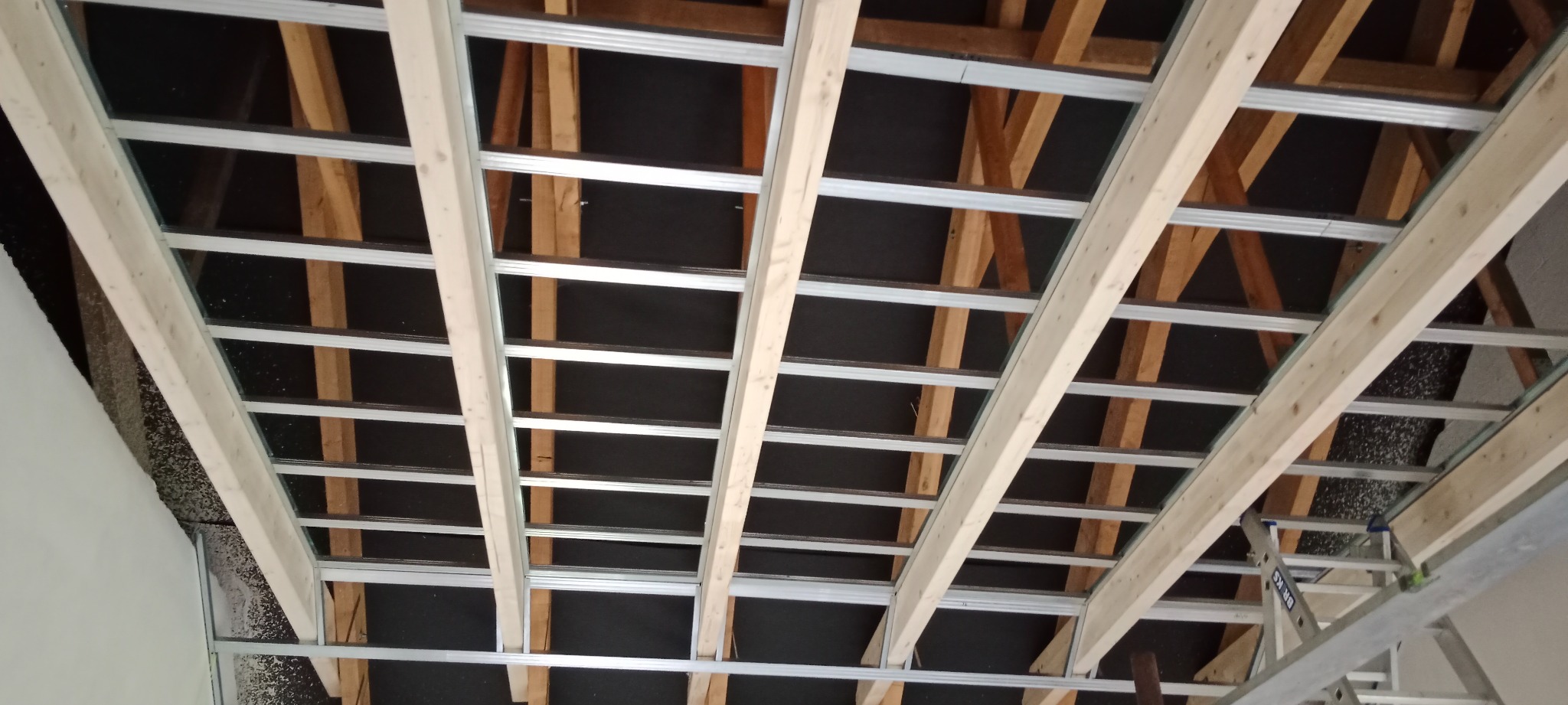 Konstrukcja sufitu podwieszanego z płyt kartonowo-gipsowych, widoczne profile metalowe i drewniane elementy więźby dachowej, czarna izolacja, drabina w rogu.