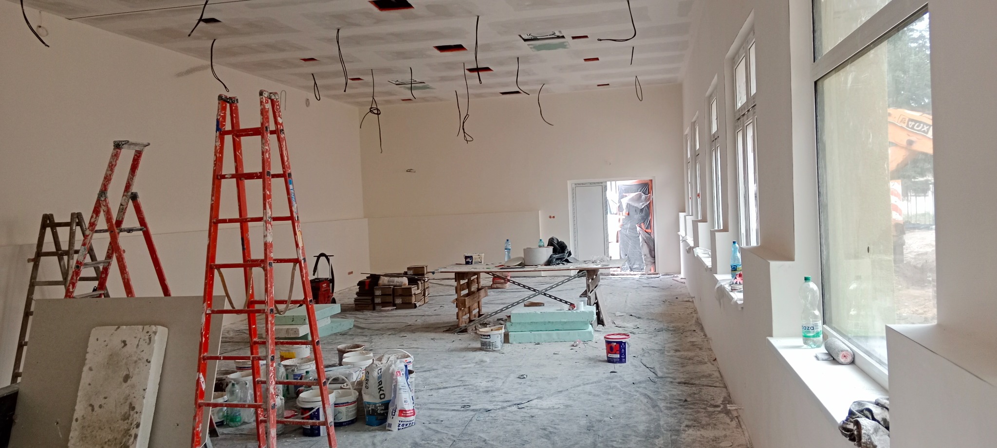 Wnętrze w trakcie prac wykończeniowych: widoczne drabiny, płyty kartonowo-gipsowe, kable zwisające z sufitu, narzędzia i materiały budowlane, okna z widokiem na plac budowy.