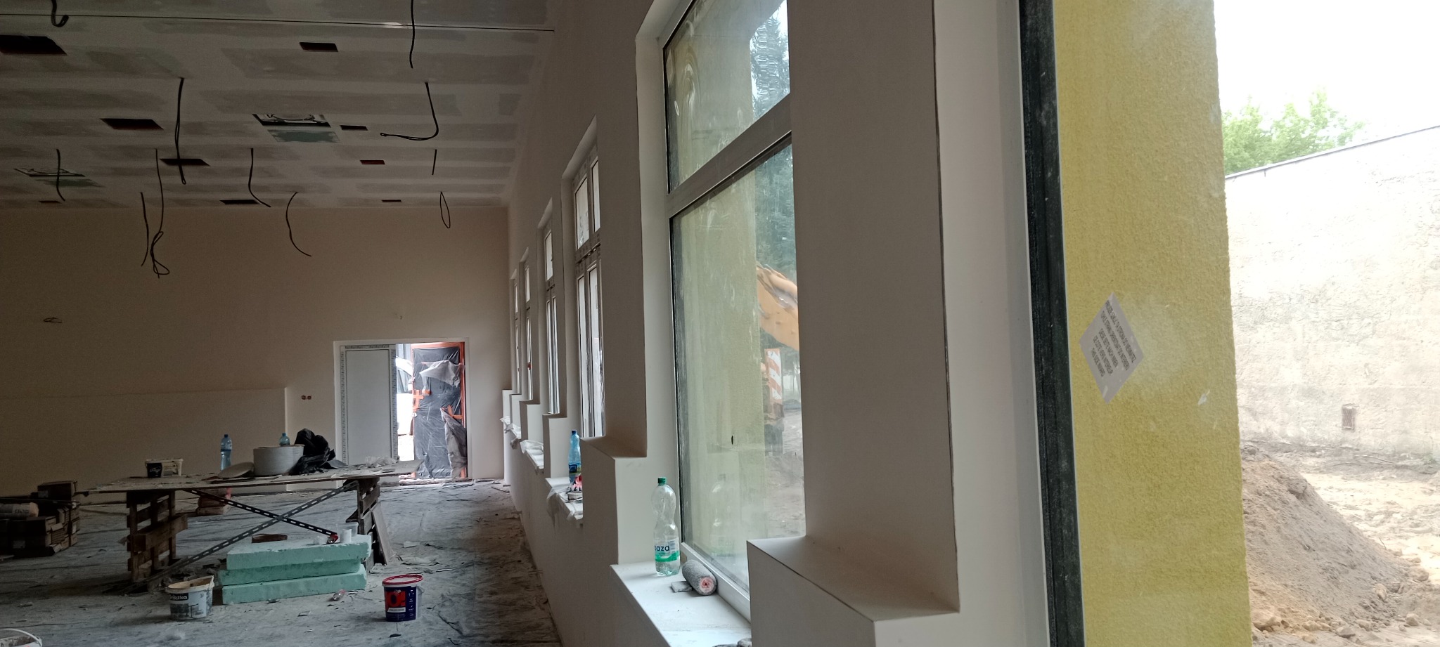 Wnętrze w trakcie prac wykończeniowych: surowy sufit z widocznymi przewodami, ściany przygotowane pod malowanie, okna z białymi ramami i widokiem na zewnątrz, narzędzia i materiały budowlane...