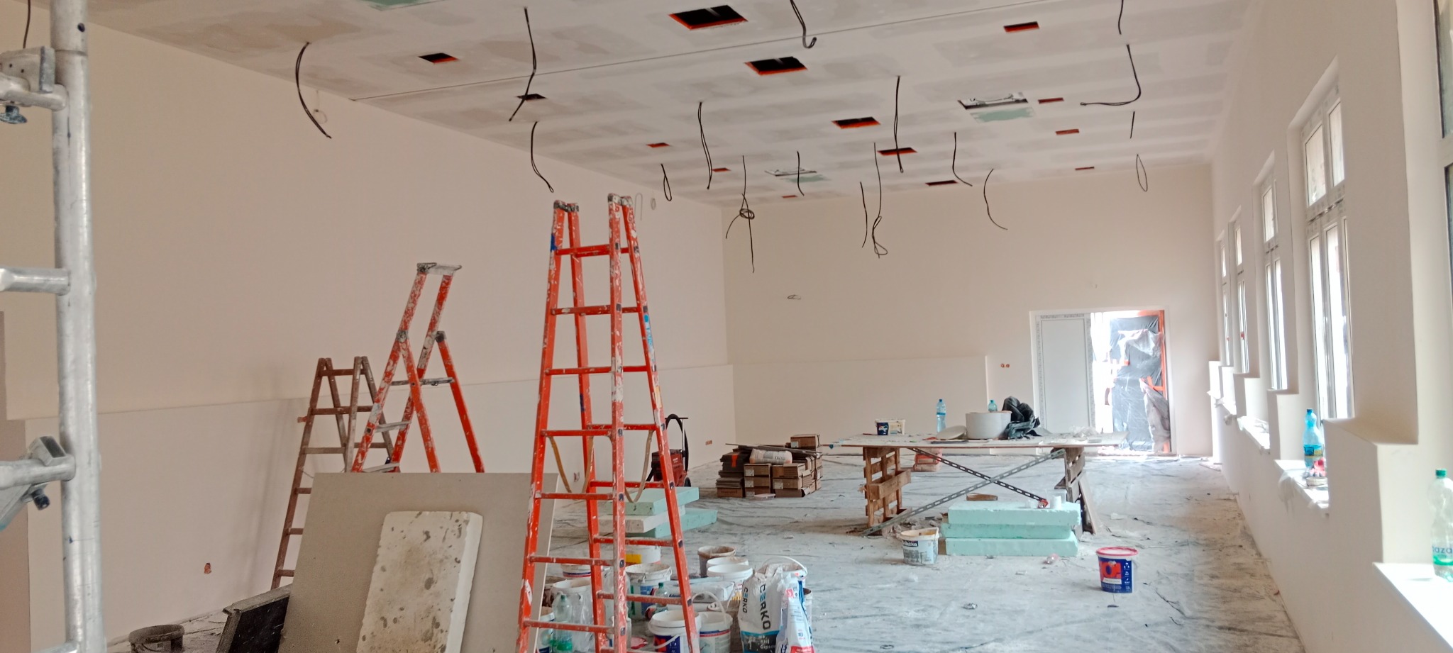 Wnętrze w trakcie prac: sufit z płyt kartonowo-gipsowych z wystającymi kablami, świeżo pomalowane ściany, drabiny, wiadra z farbą i narzędzia malarskie na podłodze zabezpieczonej folią.