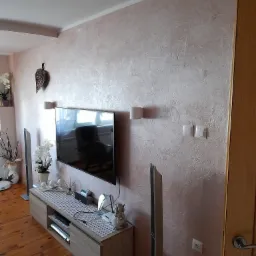 Ściana w salonie wykończona dekoracyjnym tynkiem w odcieniach różu, z widoczną fakturą i delikatnym połyskiem, oświetlona przez dwa kinkiety; na ścianie wisi telewizor i dekoracja w kształcie liścia.