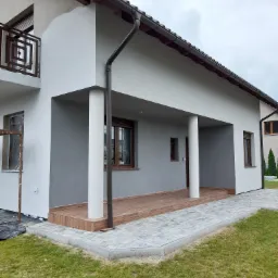 Dwukondygnacyjny dom z szarą elewacją, brązowymi oknami i drewnianym podestem werandy, częściowo ocieplony, z widocznym rusztowaniem i folią ochronną przy oknie.