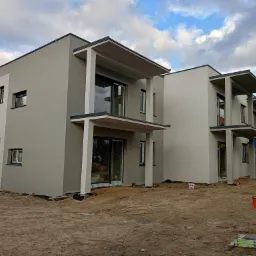 Dwa nowoczesne, dwukondygnacyjne domy w trakcie wykańczania elewacji zewnętrznej, z widocznymi puszkami farb i drabiną na placu budowy, na tle pochmurnego nieba.