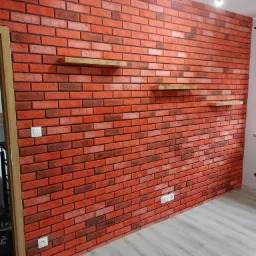 Ściana z czerwonej cegły dekoracyjnej z trzema drewnianymi półkami, widoczny włącznik światła i gniazdka elektryczne, szara podłoga i nowoczesna lampa sufitowa.