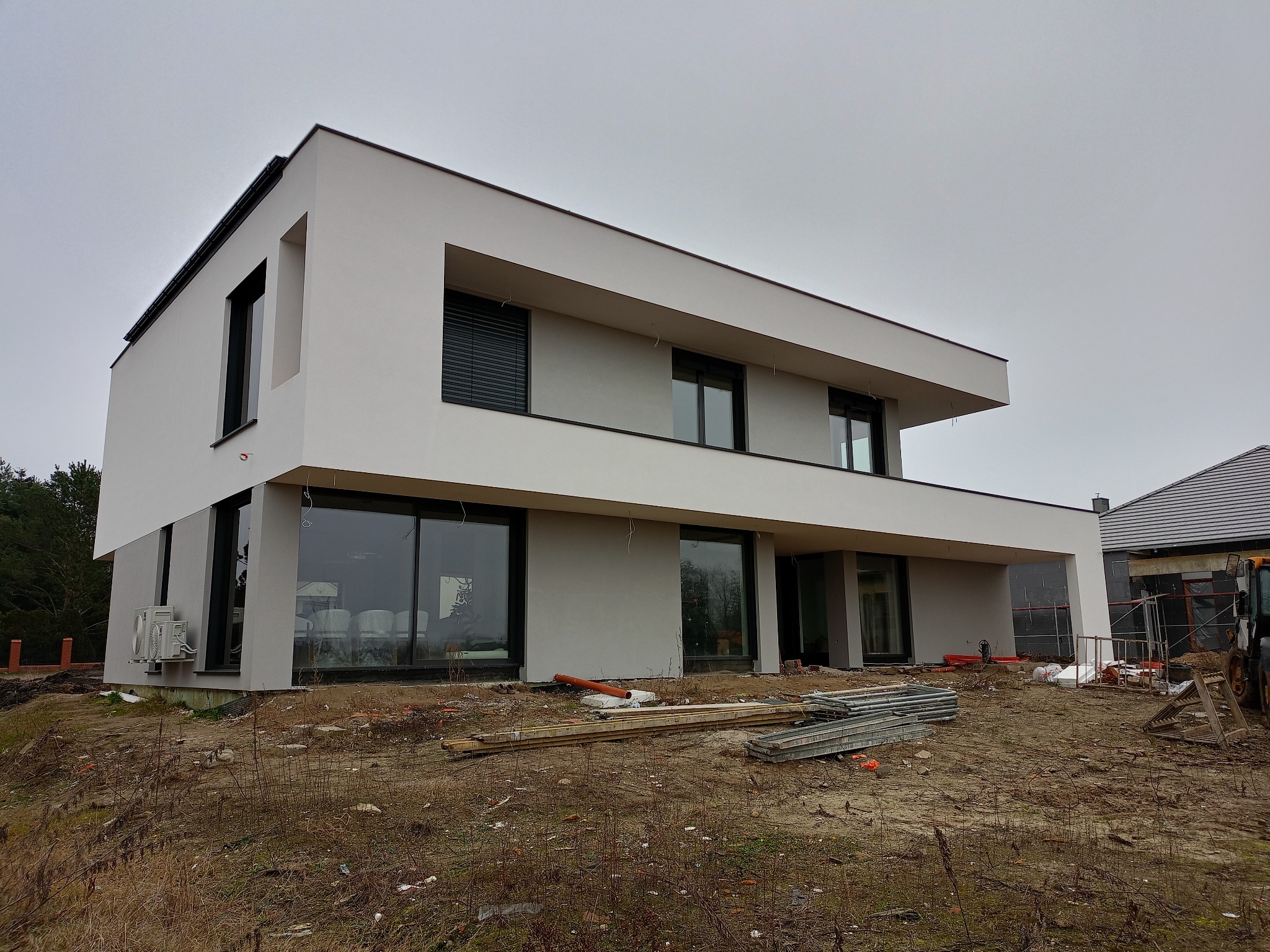 Nowoczesny, dwukondygnacyjny dom z płaskim dachem w trakcie wykończenia elewacji. Widoczne duże okna, szare ściany i elementy rusztowania. Teren wokół domu niezagospodarowany.
