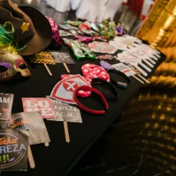 Różnorodne rekwizyty do zdjęć: kolorowe peruki, maski karnawałowe, kapelusz kowbojski, tabliczki z napisami 'Kocham Ja' i 'Taka Impreza' rozłożone na czarnym stole, złote tło