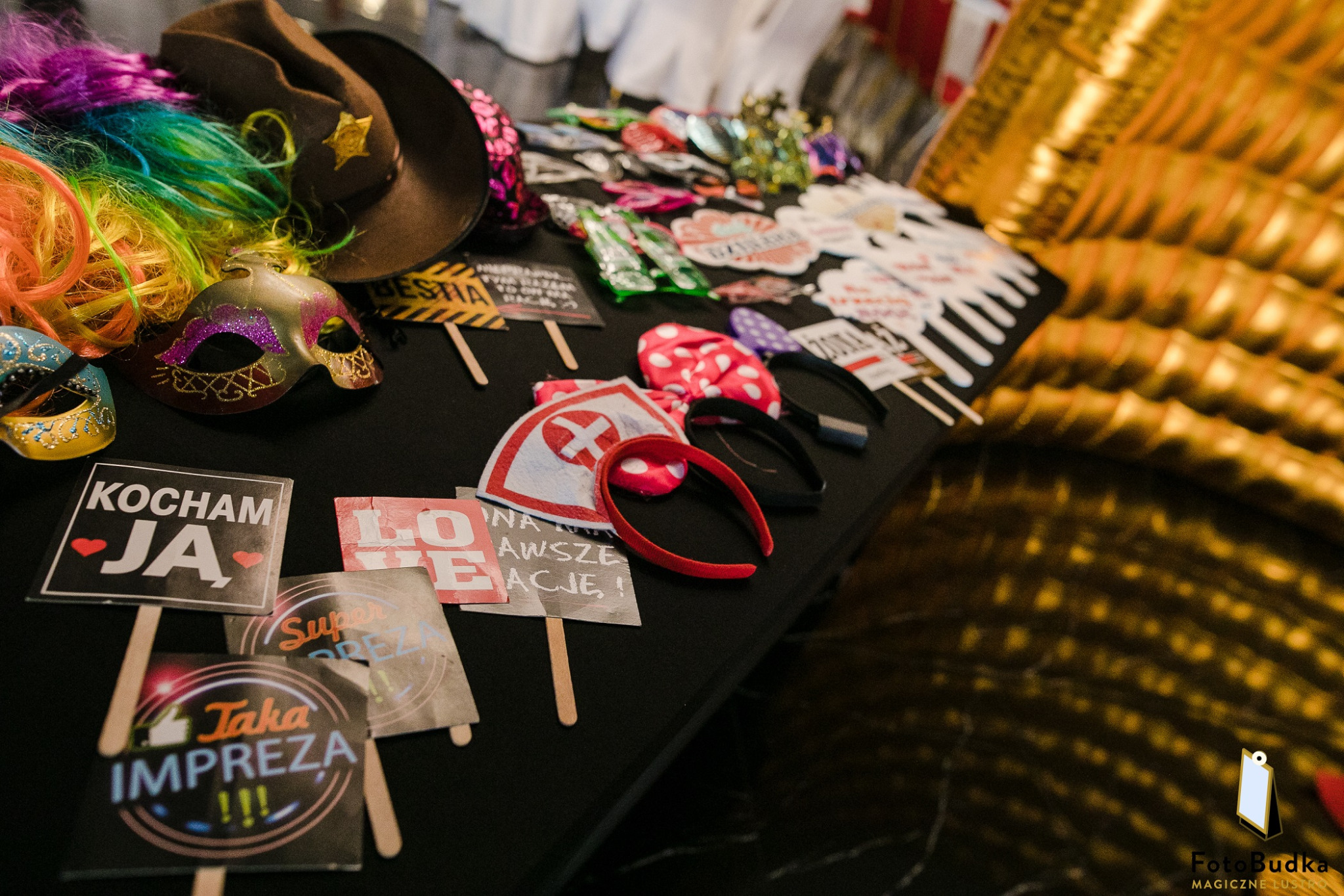 Różnorodne rekwizyty do zdjęć: kolorowe peruki, maski karnawałowe, kapelusz kowbojski, tabliczki z napisami 'Kocham Ja' i 'Taka Impreza' rozłożone na czarnym stole, złote tło