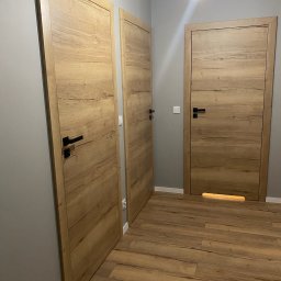 Sonder plus Denys Nutrikhin - Trzy drewniane drzwi wewnętrzne z czarnymi klamkami, zamontowane w szarym korytarzu z panelami podłogowymi w odcieniach brązu; jedno z drzwi posiada podświetlenie dolne.