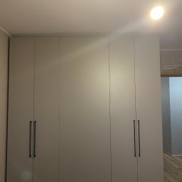 Sonder plus Denys Nutrikhin - Zamknięta, czterodrzwiowa szafa wnękowa z długimi, pionowymi uchwytami na tle jasnego pokoju z drewnianą podłogą i wiszącą żarówką.