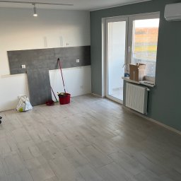 Sonder plus Denys Nutrikhin - Wnętrze w trakcie remontu: częściowo położone szare płytki na ścianie, jasne panele podłogowe imitujące drewno, wiadro i narzędzia malarskie, kartonowe pudła przy oknie balkonowym, grzejnik.