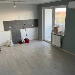 Wnętrze w trakcie remontu: częściowo położone szare płytki na ścianie, jasne panele podłogowe imitujące drewno, wiadro i narzędzia malarskie, kartonowe pudła przy oknie balkonowym, grzejnik.