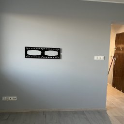 Sonder plus Denys Nutrikhin - Szara ściana z zamontowanym uchwytem do telewizora, widoczne gniazdka elektryczne i włączniki światła, podłoga wyłożona panelami, w tle otwarte drzwi do innego pomieszczenia.