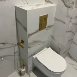 Nowoczesna toaleta podwieszana z zabudowaną spłuczką, wykończona płytkami imitującymi marmur z akcentami w kolorze złotym.