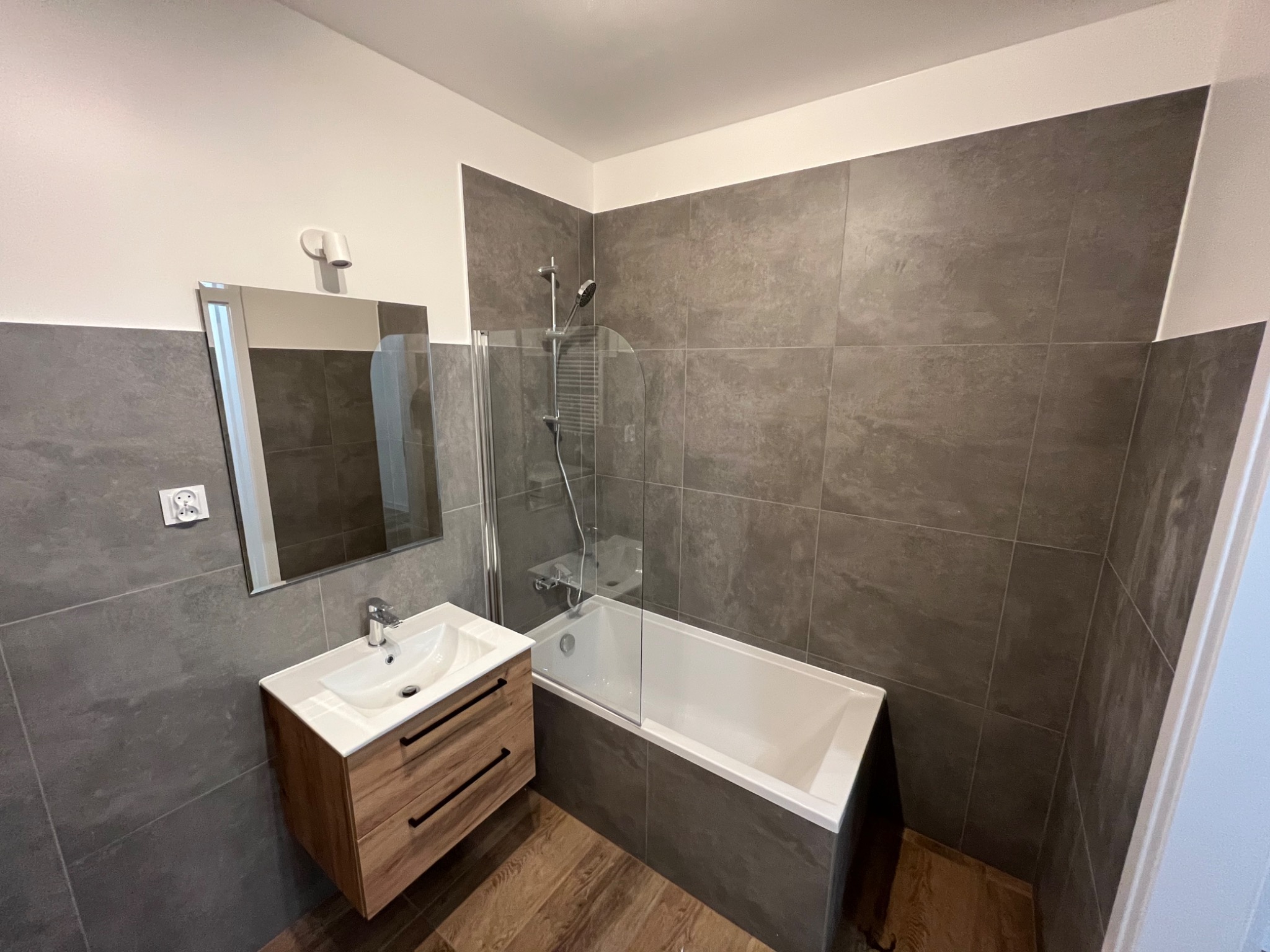 Nowoczesna łazienka z szarymi płytkami imitującymi kamień, drewnianą szafką pod umywalką z czarnymi uchwytami, białą wanną z parawanem i drewnopodobną podłogą.