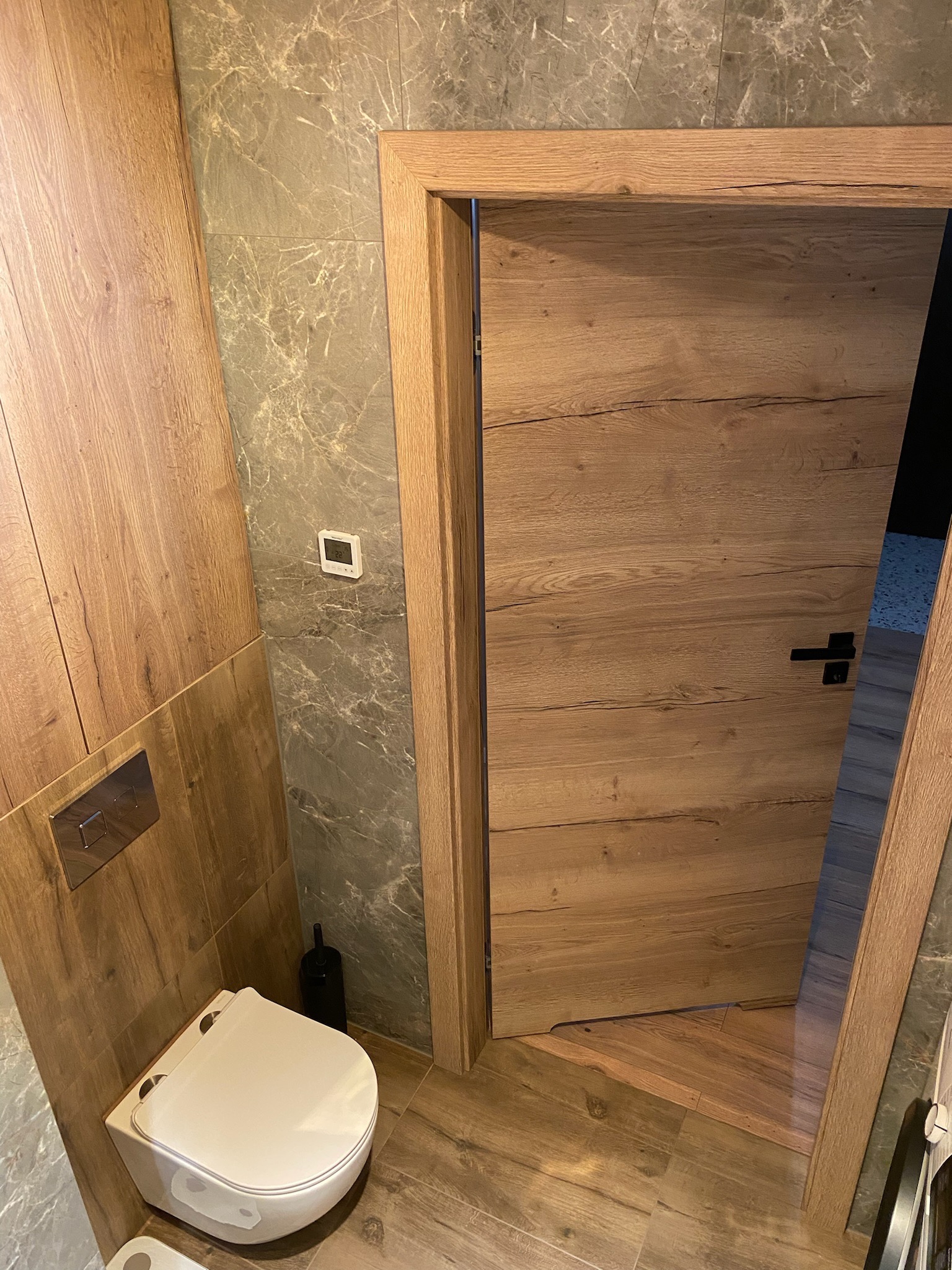 Nowoczesna łazienka z drewnianą zabudową, marmurowymi płytkami i podwieszaną toaletą, widok na otwarte drzwi z drewnianą ościeżnicą.