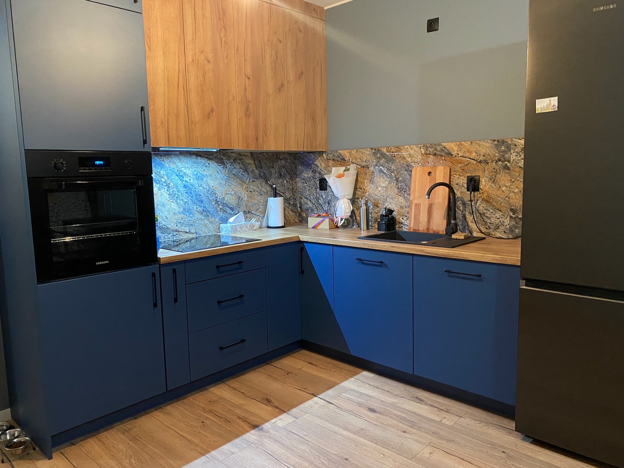 Nowoczesna kuchnia z granatowymi szafkami, drewnianymi górnymi szafkami, blatem z drewnopodobnego laminatu i marmurowym backsplashiem. Widoczny piekarnik Samsung, lodówka i drewniana deska...