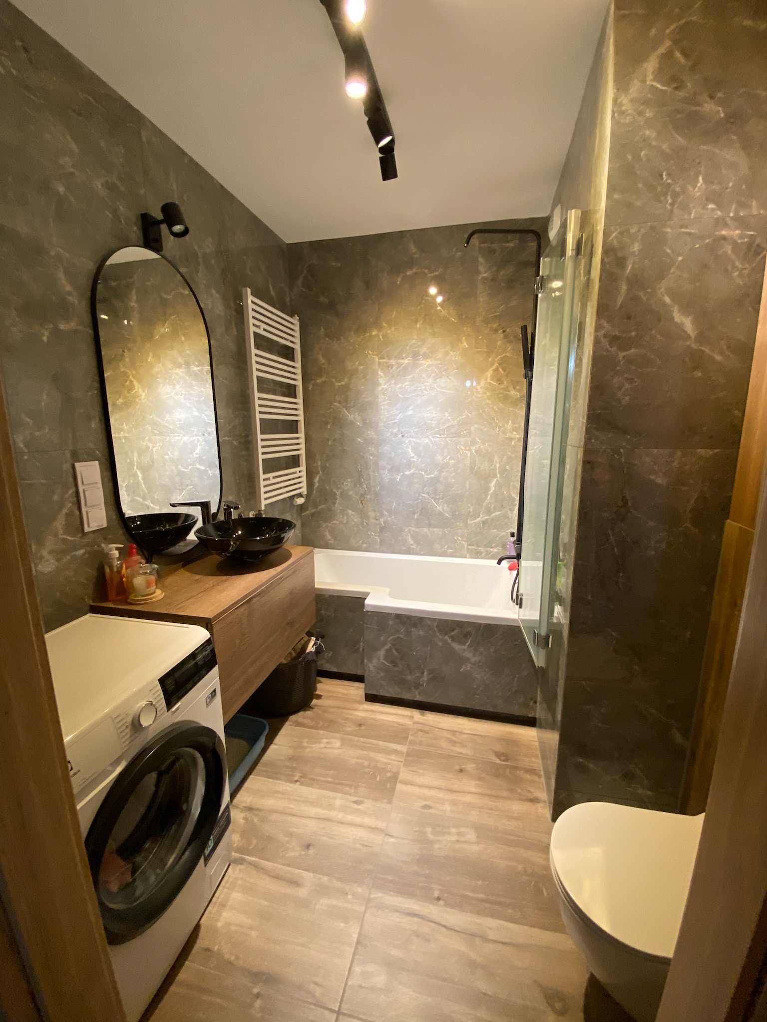 Nowoczesna łazienka z marmurowymi płytkami, dwoma czarnymi umywalkami na drewnianej szafce, pralką i kabiną prysznicową z wanną.