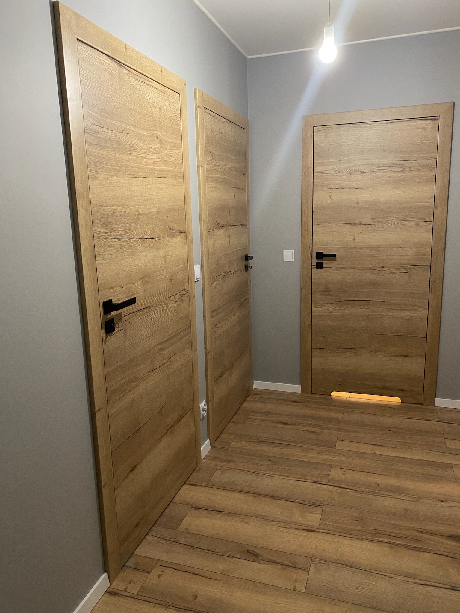 Trzy drewniane drzwi wewnętrzne z czarnymi klamkami, zamontowane w szarym korytarzu z panelami podłogowymi w odcieniach brązu; jedno z drzwi posiada podświetlenie dolne.