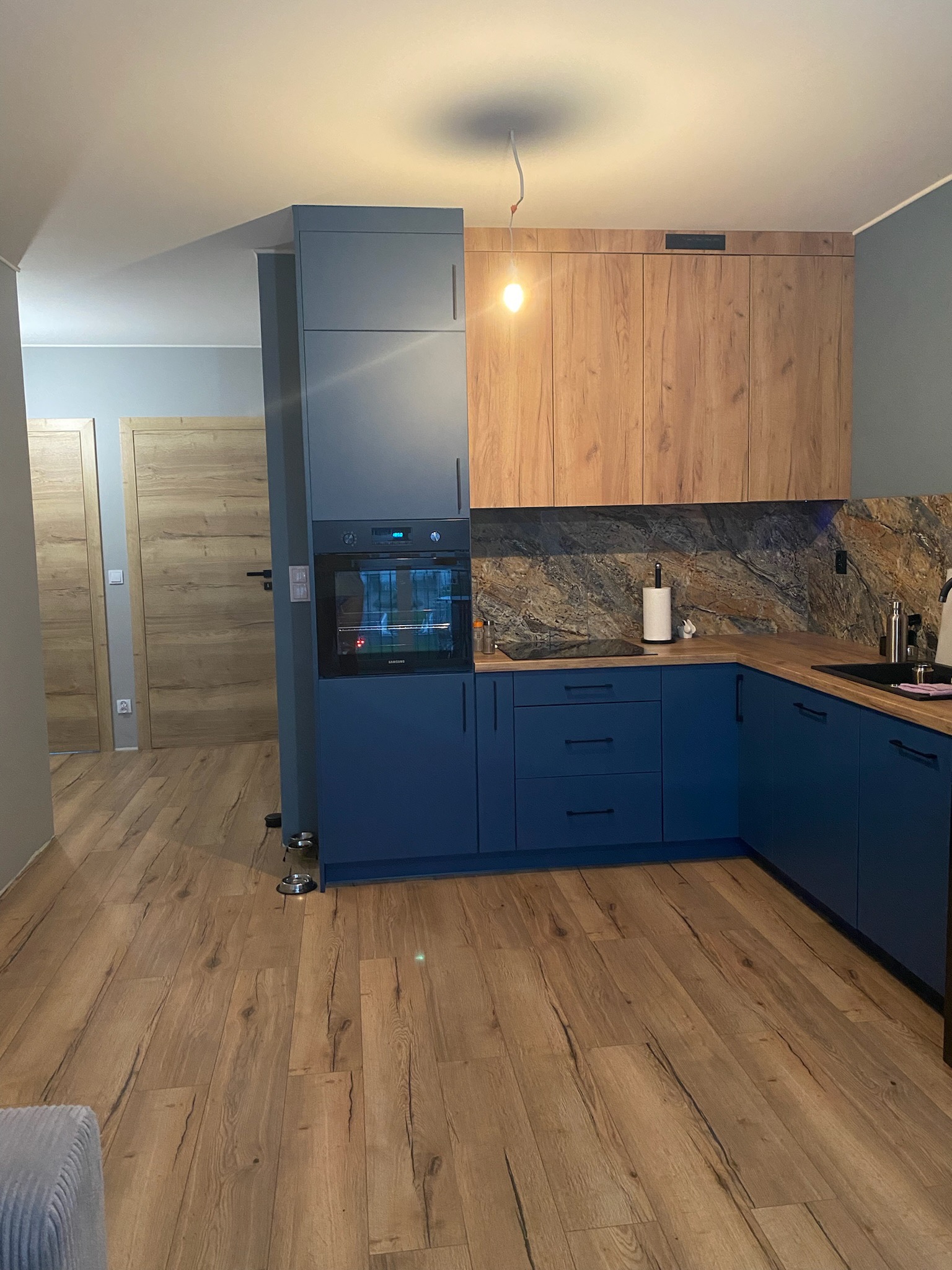 Nowoczesna kuchnia z niebieskimi szafkami, drewnianymi blatami i marmurowym panelem nad blatem, z widoczną zabudowaną piekarnikiem i drewnianą podłogą.