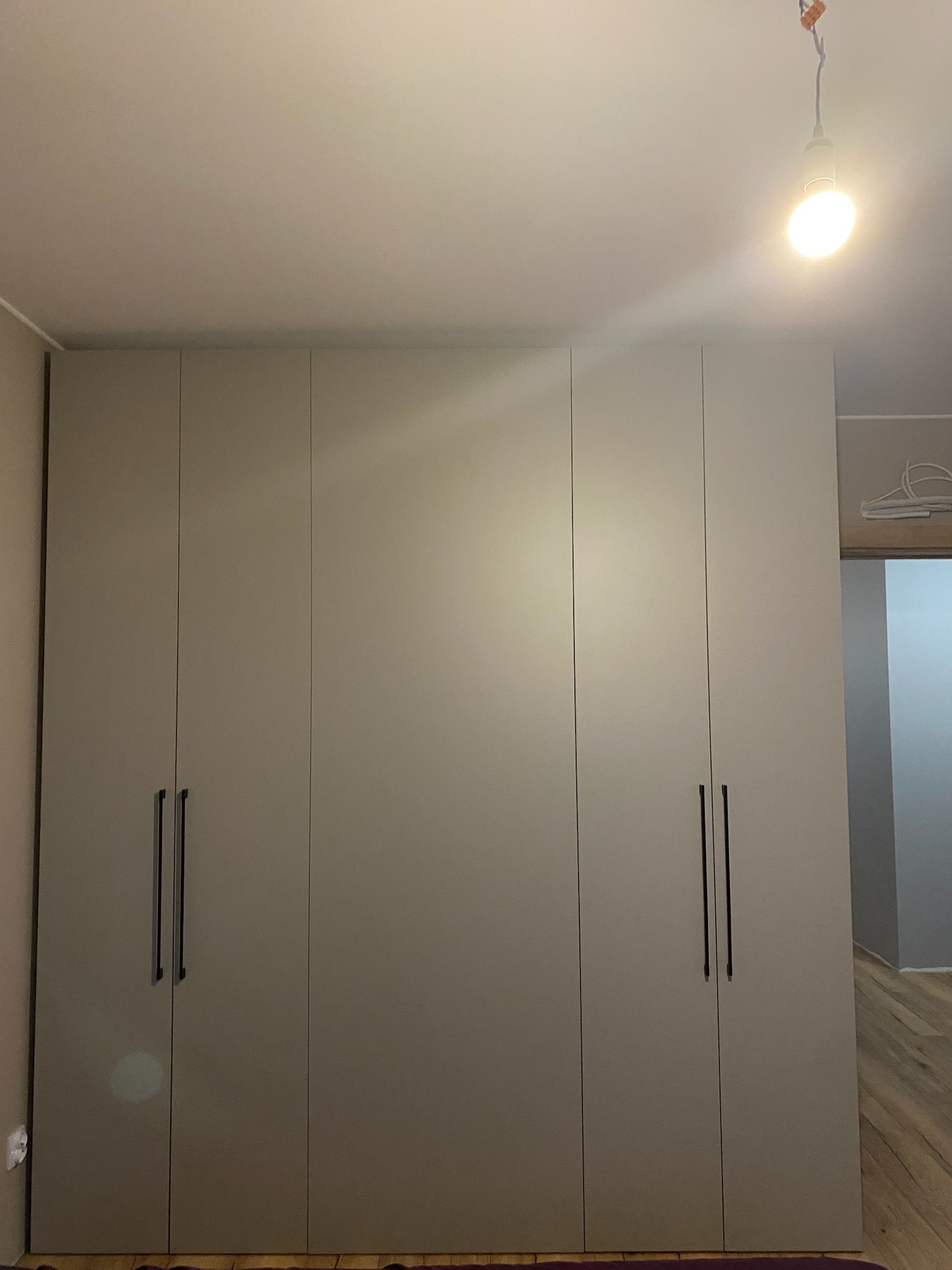 Zamknięta, czterodrzwiowa szafa wnękowa z długimi, pionowymi uchwytami na tle jasnego pokoju z drewnianą podłogą i wiszącą żarówką.