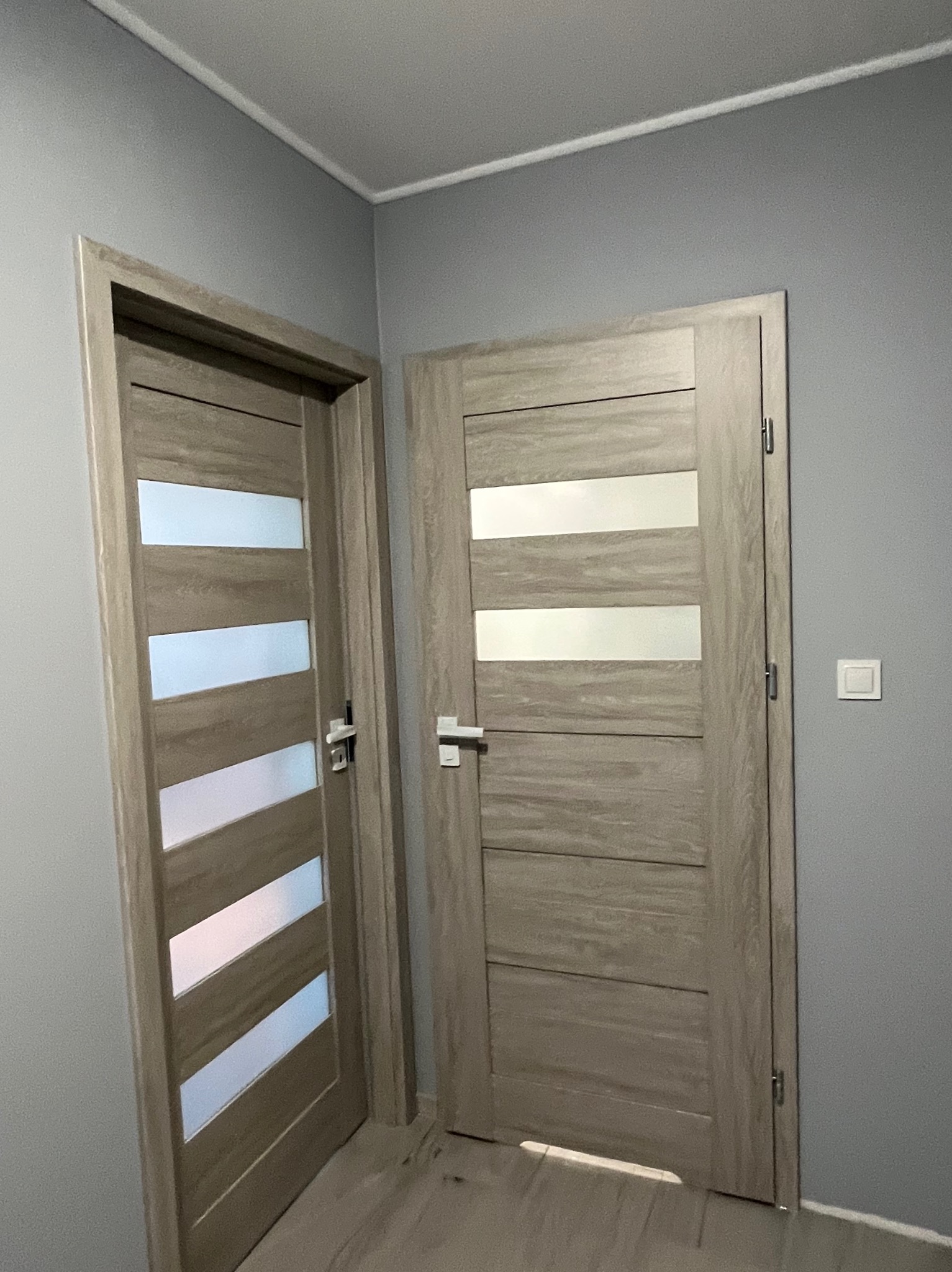 Dwa zamontowane, wewnętrzne drzwi z poziomymi przeszkleniami, w odcieniach szarości, na tle szarej ściany z włącznikiem.