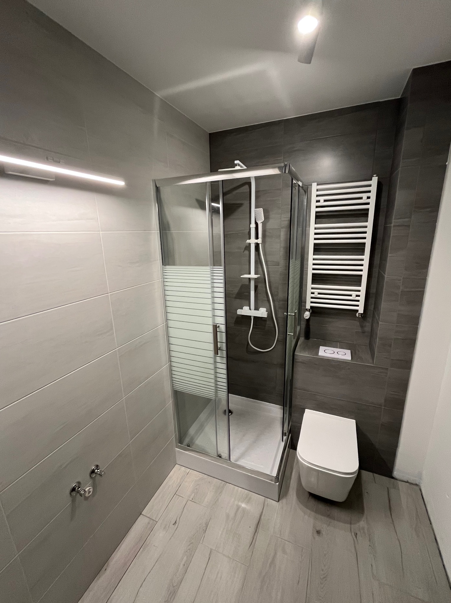 Nowoczesna łazienka z szarą glazurą, kabiną prysznicową z częściowo matową szybą, wiszącą toaletą i grzejnikiem drabinkowym.