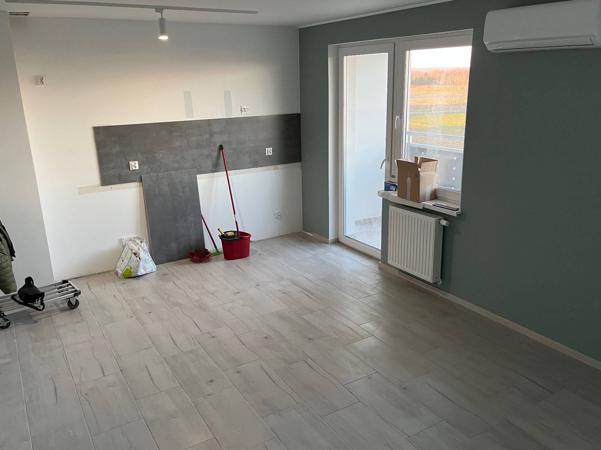 Wnętrze w trakcie remontu: częściowo położone szare płytki na ścianie, jasne panele podłogowe imitujące drewno, wiadro i narzędzia malarskie, kartonowe pudła przy oknie balkonowym, grzejnik.