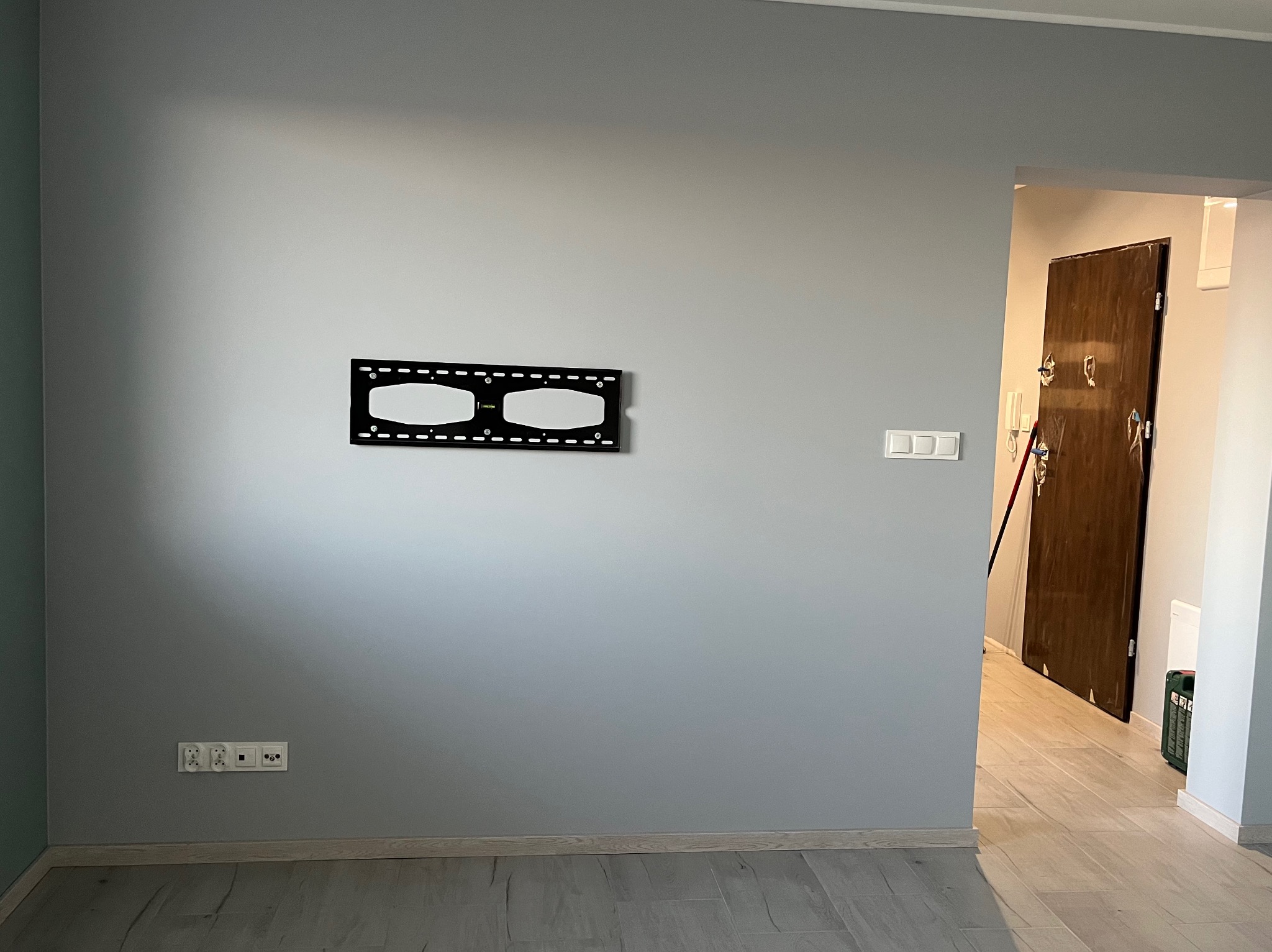 Szara ściana z zamontowanym uchwytem do telewizora, widoczne gniazdka elektryczne i włączniki światła, podłoga wyłożona panelami, w tle otwarte drzwi do innego pomieszczenia.