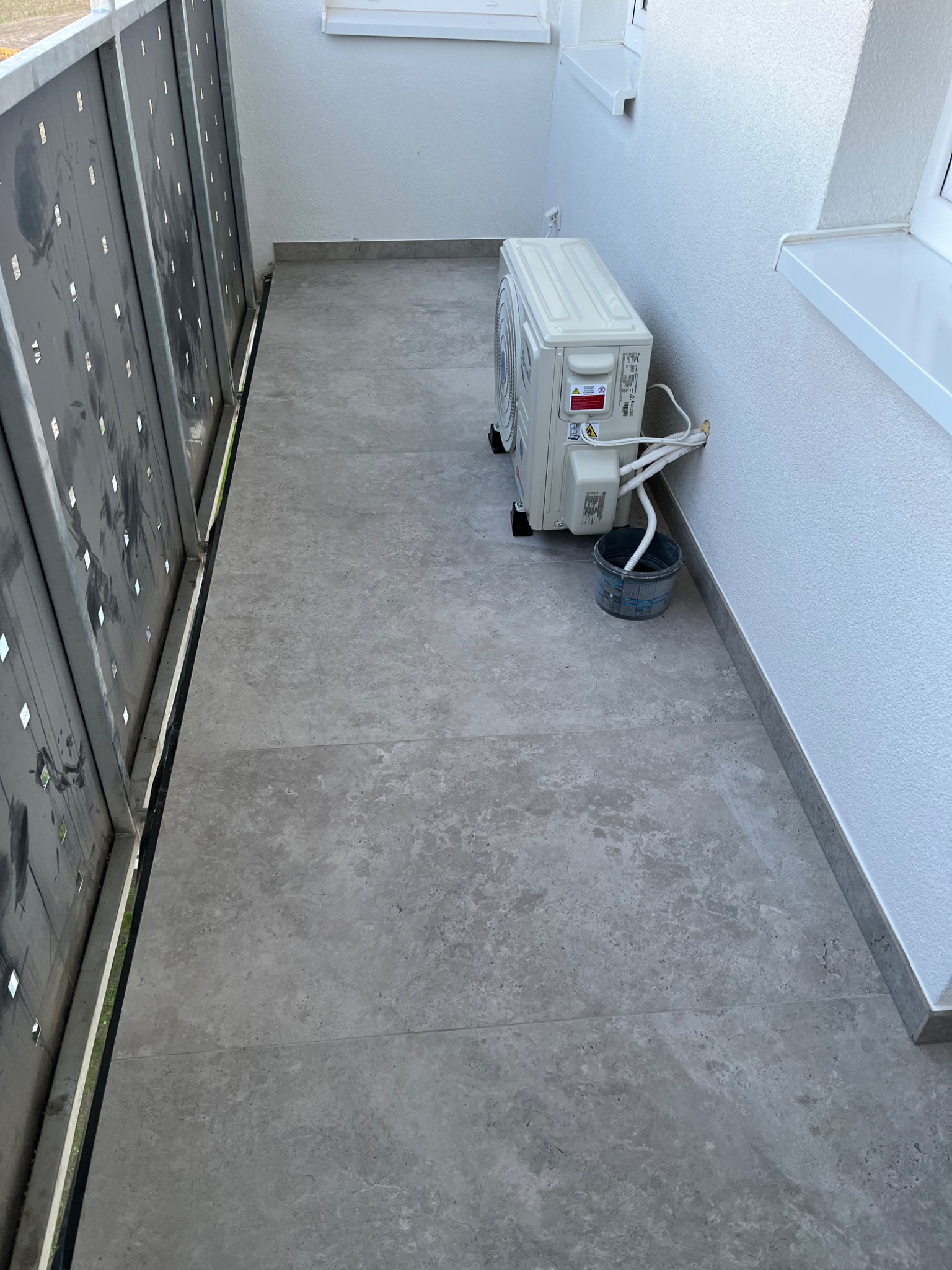 Kompaktowa jednostka zewnętrzna klimatyzacji zamontowana na balkonie z szarymi płytkami, widoczne białe rury i wiadro na skropliny.