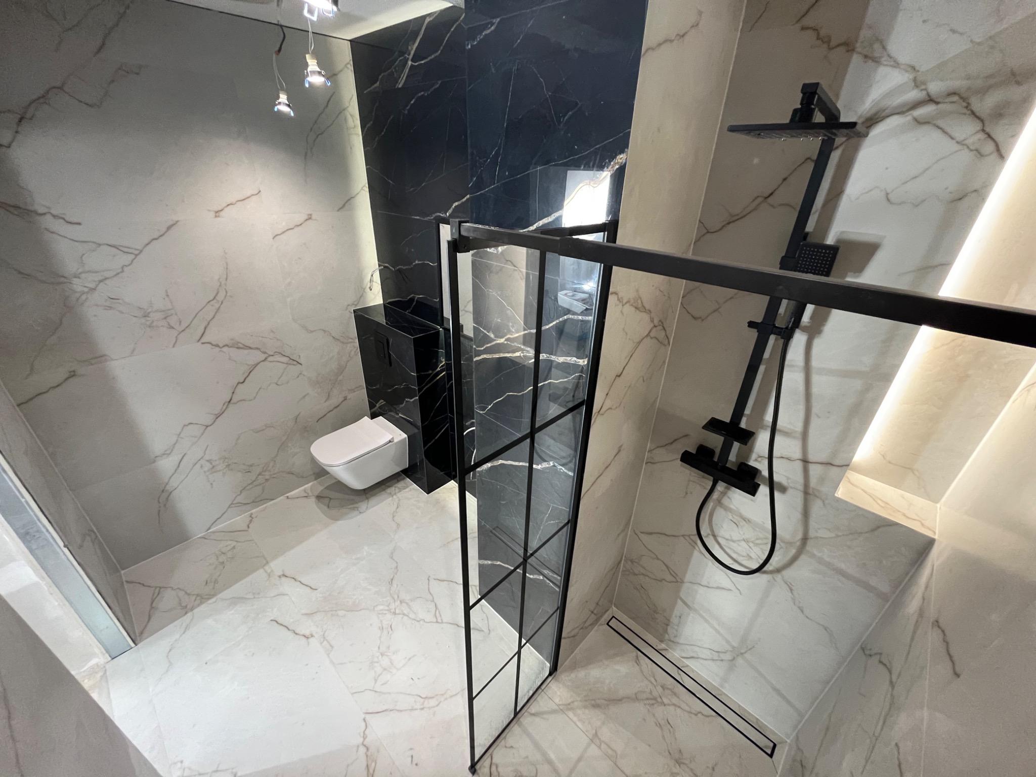Nowoczesna łazienka z czarnymi akcentami: kabina prysznicowa typu walk-in z czarną ramą, deszczownica, wisząca toaleta i marmurowe płytki w odcieniach szarości i czerni.