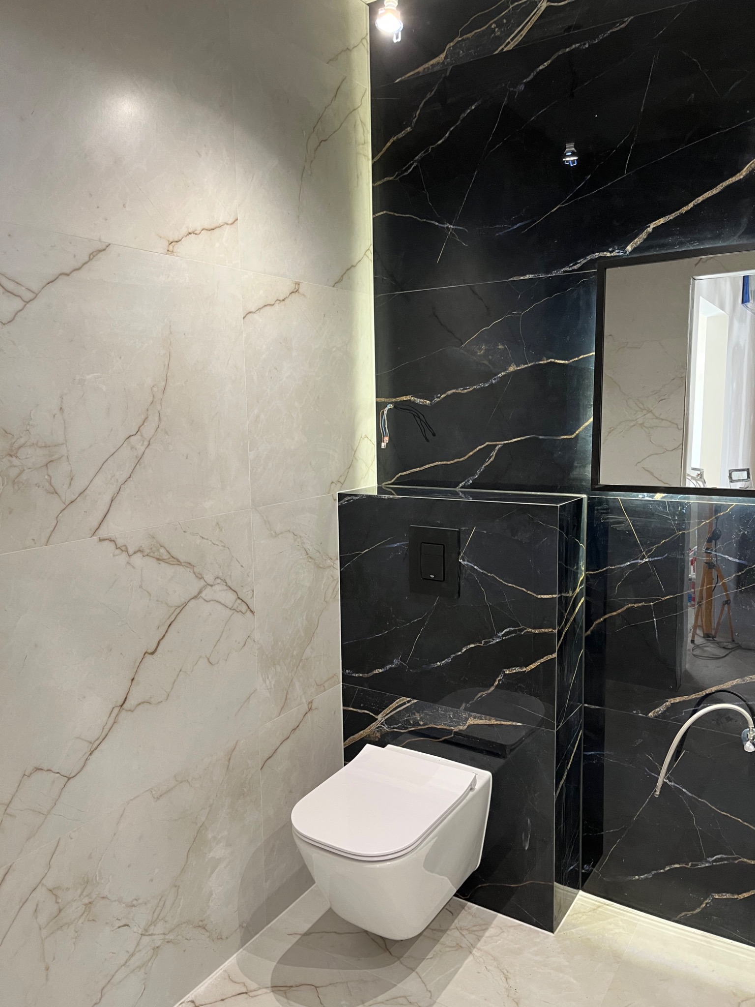 Nowoczesna łazienka we Wrocławiu z kontrastującymi płytkami: białe marmurowe i czarne z żyłami, wisząca toaleta, lustro i niedokończone podłączenie oświetlenia.