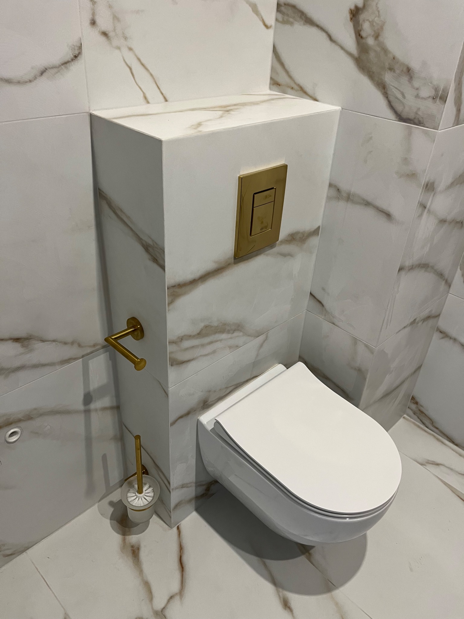 Nowoczesna toaleta podwieszana z zabudowaną spłuczką, wykończona płytkami imitującymi marmur z akcentami w kolorze złotym.