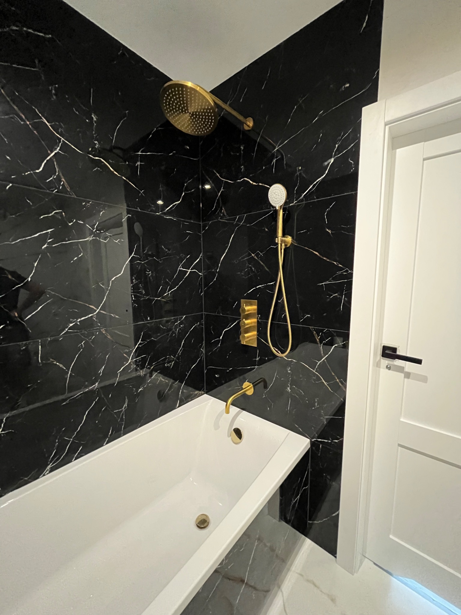 Nowoczesna łazienka z czarnymi marmurowymi płytkami, złotą armaturą i białą wanną, widok na prysznic i drzwi.