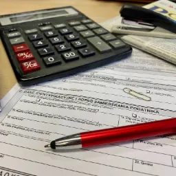 Biurko z kalkulatorem, zszywaczem, długopisem i wypełnianym formularzem podatkowym, gazeta w tle.