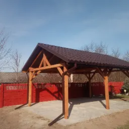 Drewniana wiata garażowa z brązowym dachem z blachodachówki, widoczna rynna, na tle czerwonego ogrodzenia i bezlistnych drzew.