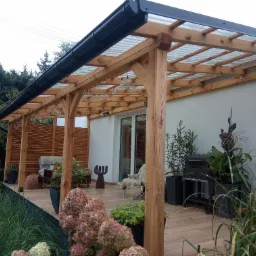 Drewniana wiata garażowa przylegająca do domu z przezroczystym zadaszeniem, czarną rynną i oświetleniem w postaci żarówek, otoczona zielenią i kwiatami w donicach na drewnianym tarasie.