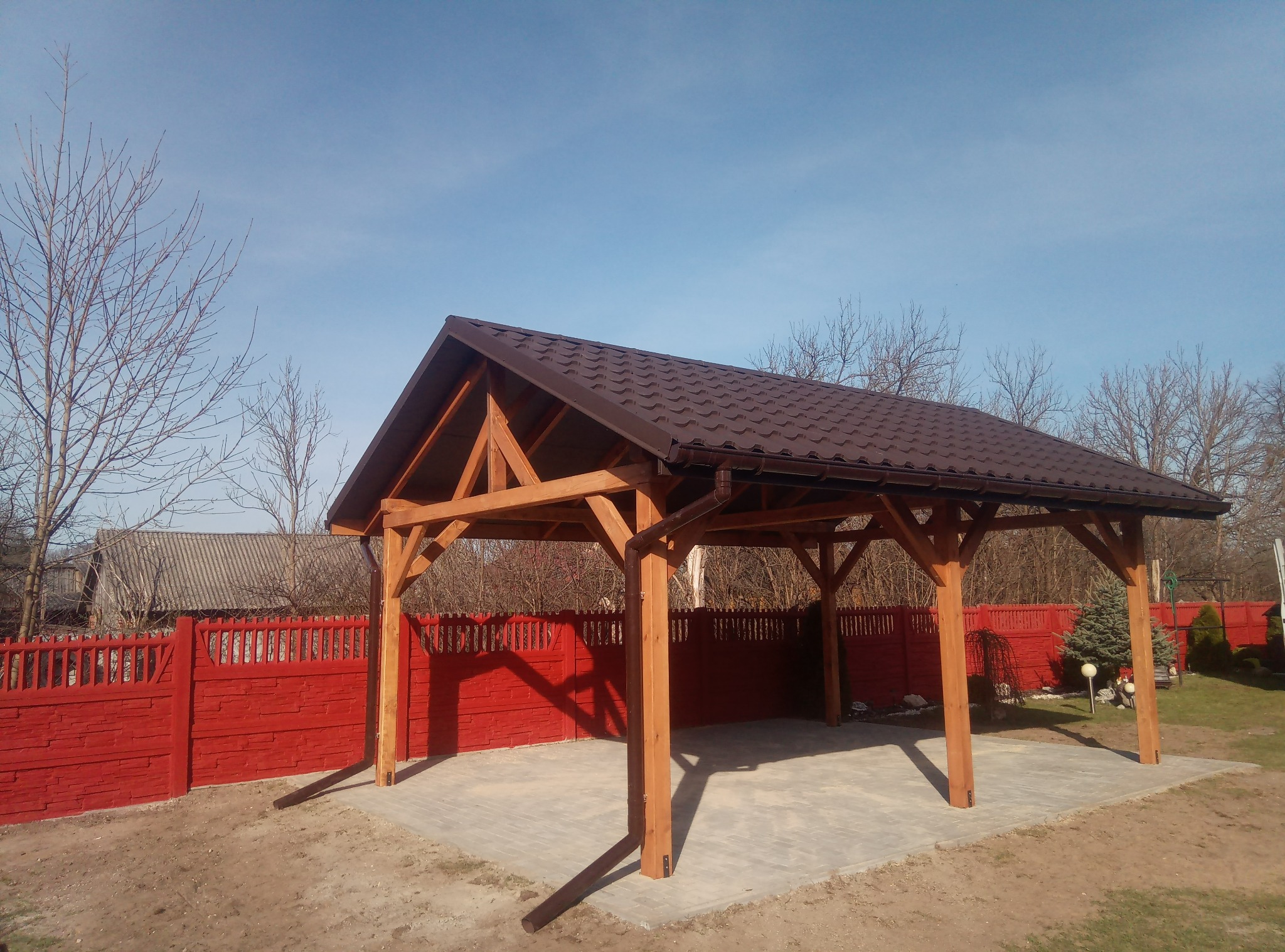 Drewniana wiata garażowa z brązowym dachem z blachodachówki, widoczna rynna, na tle czerwonego ogrodzenia i bezlistnych drzew.