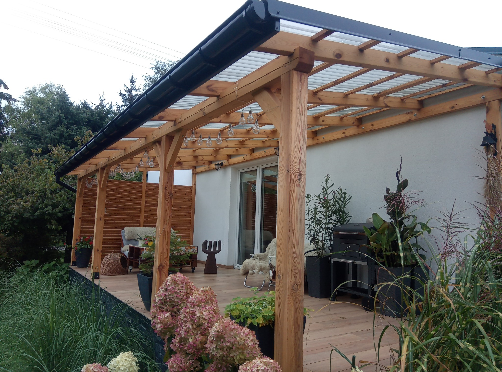 Drewniana wiata garażowa przylegająca do domu z przezroczystym zadaszeniem, czarną rynną i oświetleniem w postaci żarówek, otoczona zielenią i kwiatami w donicach na drewnianym tarasie.