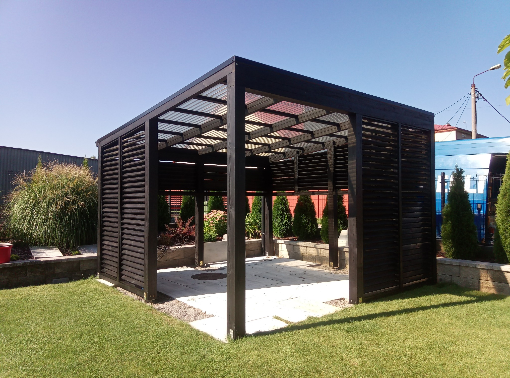Nowoczesna, ciemna altana z przezroczystym dachem, otoczona zielonym trawnikiem i dekoracyjnymi roślinami, w słoneczny dzień.